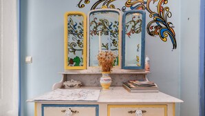 Une pièce avec un mur bleu, une porte blanche, une coiffeuse avec trois miroirs, un vase contenant des fleurs séchées et une carte posée sur le comptoir.