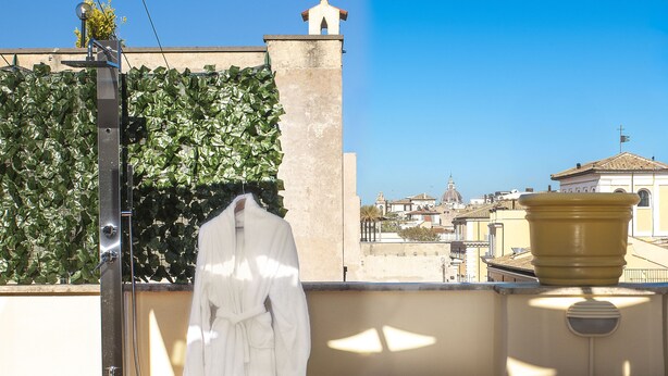 A balcony with a white bathrobe hanging, a shower, and a view of a cityscape. - Cestari Luxury Terraces (Rome)