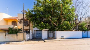Rua residencial com uma casa, portão fechado, uma árvore em frente e céu claro.
