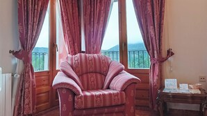 Une pièce comprenant un fauteuil confortable, une table en bois avec des documents, et une vue sur un paysage montagneux grâce à de grandes fenêtres ornées de rideaux à motifs.