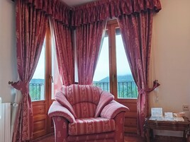 Una habitación con un sillón mullido, una mesa de madera con documentos y vistas a un paisaje montañoso a través de ventanas amplias con cortinas estampadas.