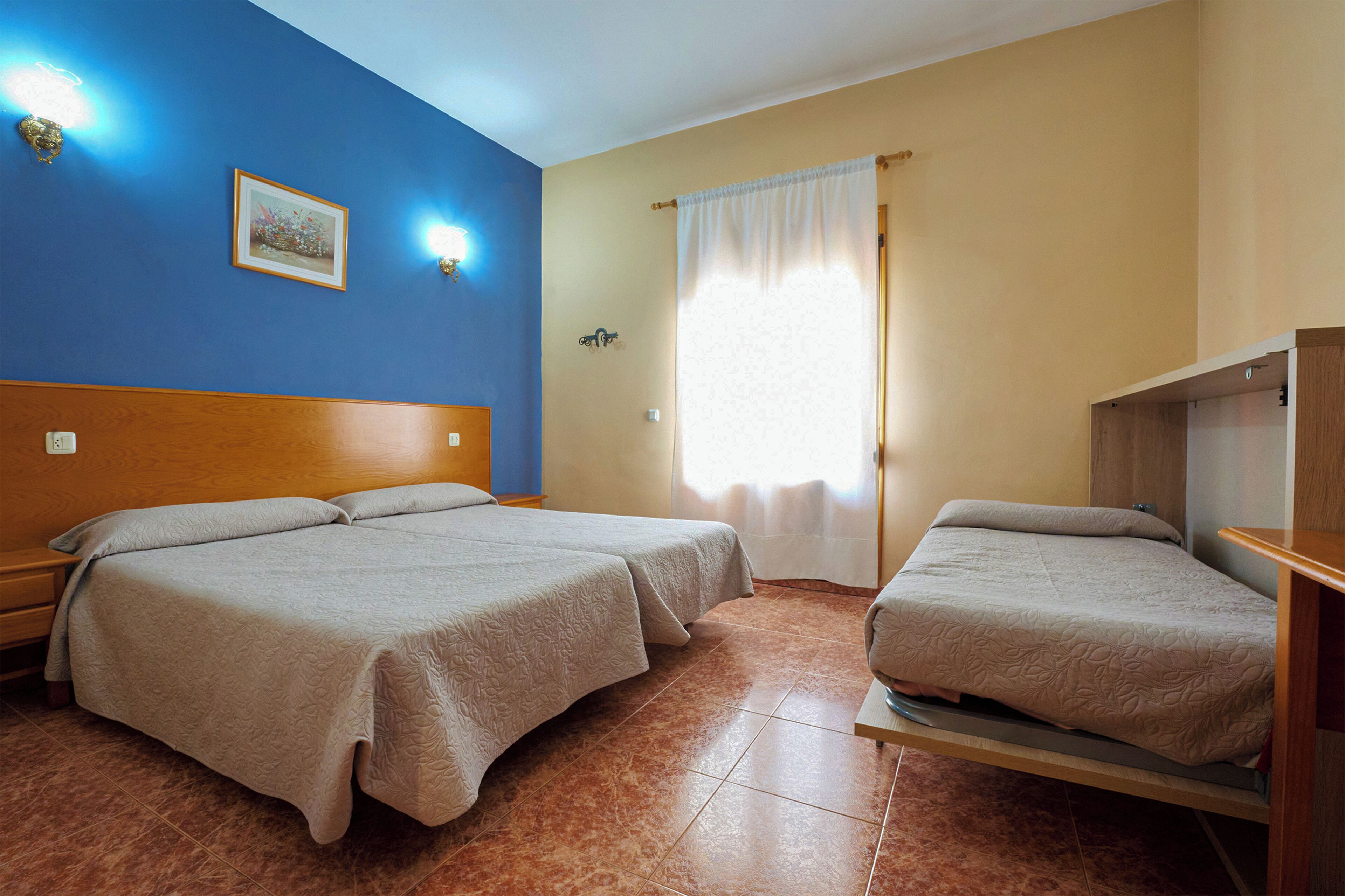 Habitación de hotel con dos camas, cabecero de madera, mesita de noche, lámpara de pared y ventana con cortinas.
