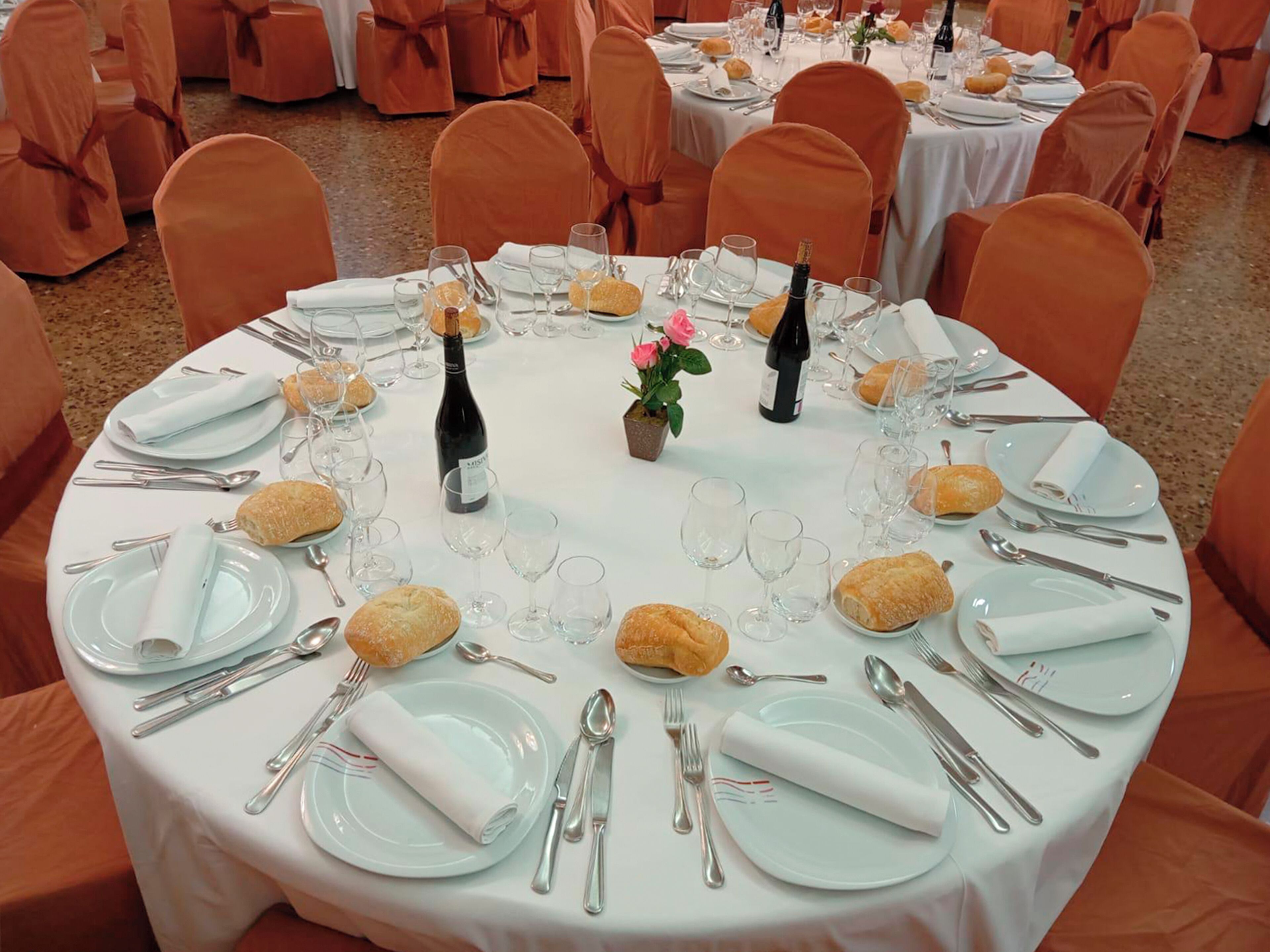 A round dining table set with white tablecloths, silverware, plates, glasses, and bread rolls.