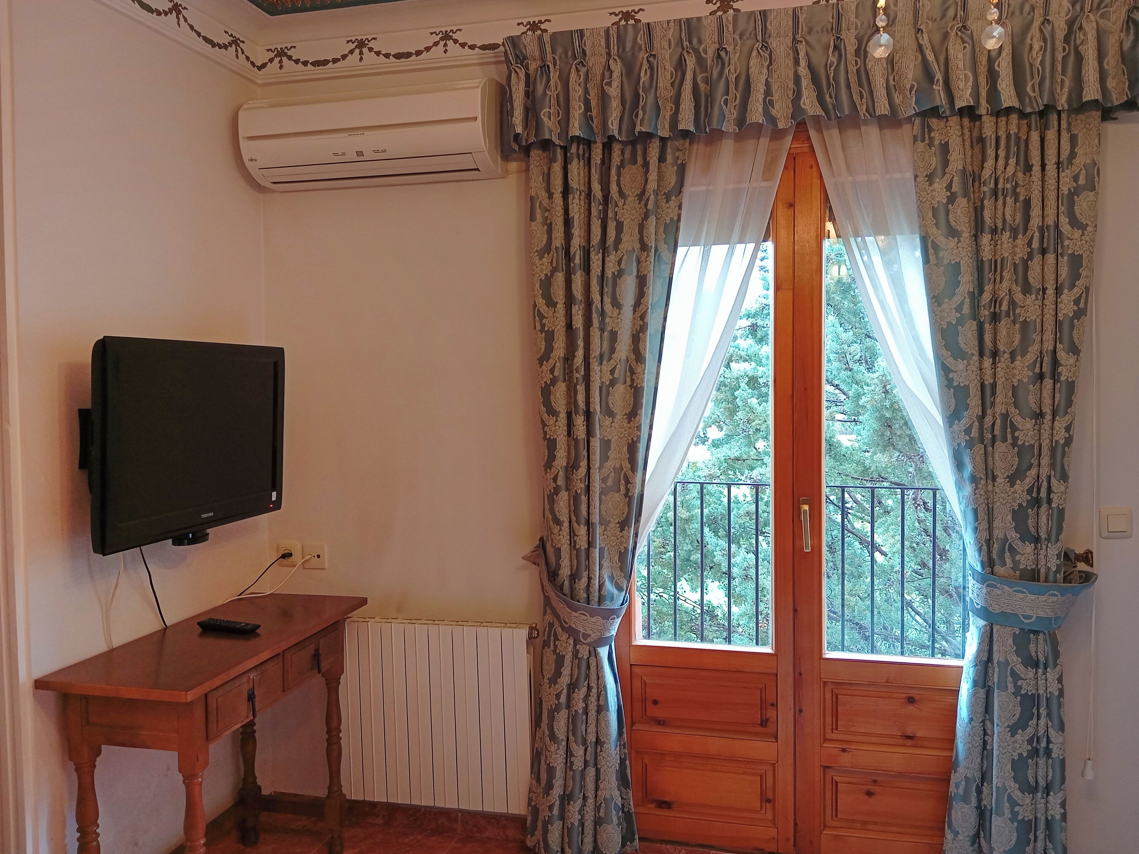 Una habitación con una televisión sobre un escritorio de madera, una ventana con cortinas estampadas y un equipo de aire acondicionado en la pared.