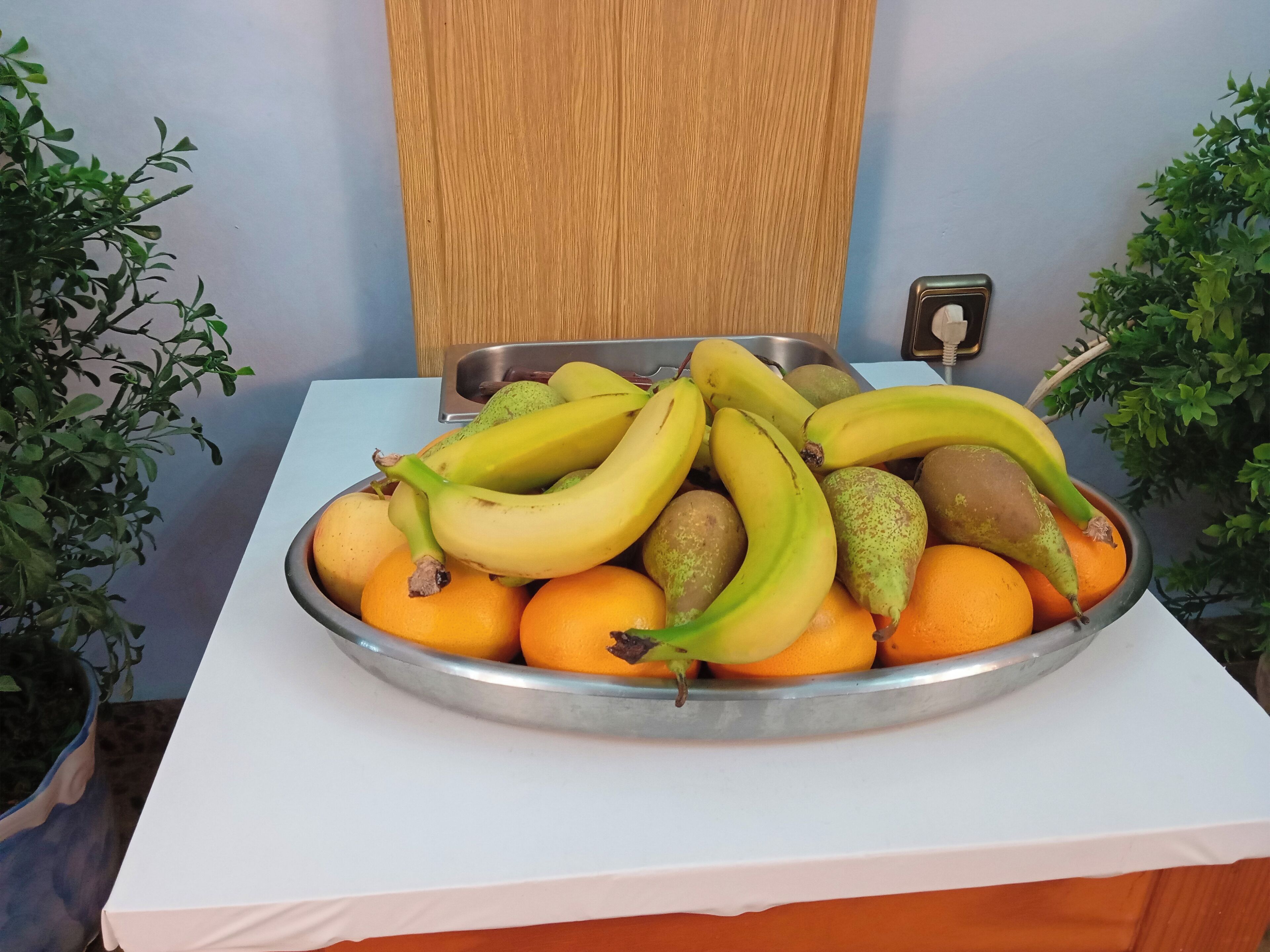 Un panier de fruits est posé sur une table recouverte d’une nappe blanche, et des plantes en pot l’entourent.