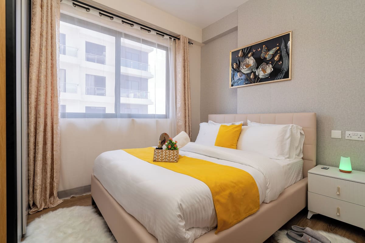 Una cama bien tendida con ropa de cama blanca y amarilla, una mesita de noche con una lámpara, y un cuadro en la pared.