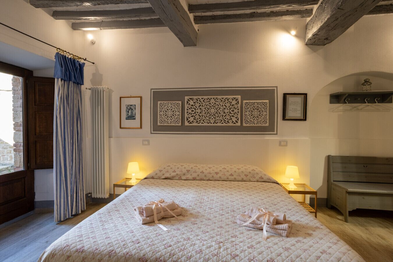 Quarto com uma cama grande, duas luminárias de cabeceira, um banco de madeira e uma janela com cortinas.