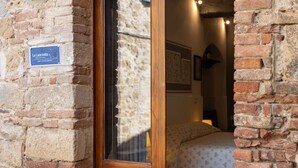 A brick building entrance with a wooden door, a sign reading "La Lanciotta," and a bed with a floral bedspread visible inside. - Camera Lanciotta - Monticchiello (Monticchiello)