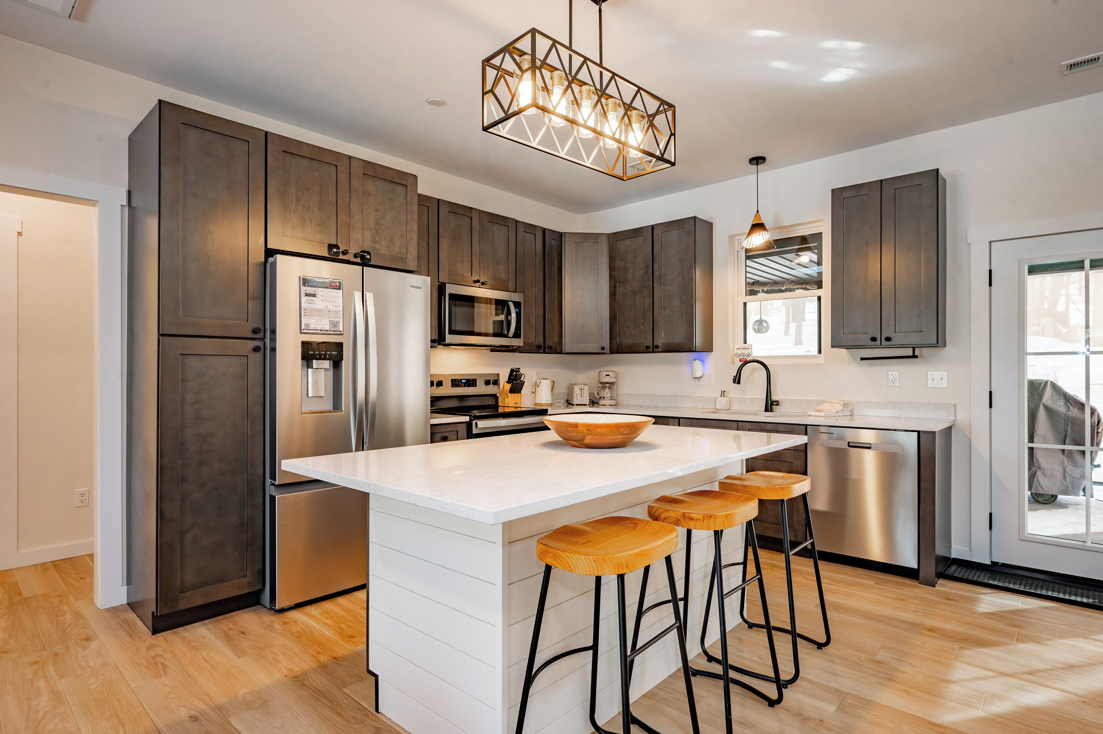 A modern kitchen with dark wooden cabinets, stainless steel appliances, a white island with bar stools, and pendant lighting.