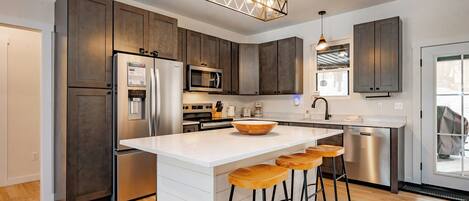 A modern kitchen with dark wooden cabinets, stainless steel appliances, a white island with bar stools, and pendant lighting.