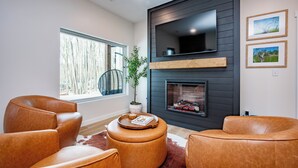 Een moderne woonkamer met leren banken, een flatscreen televisie, een open haard en een groot raam met uitzicht op bomen.