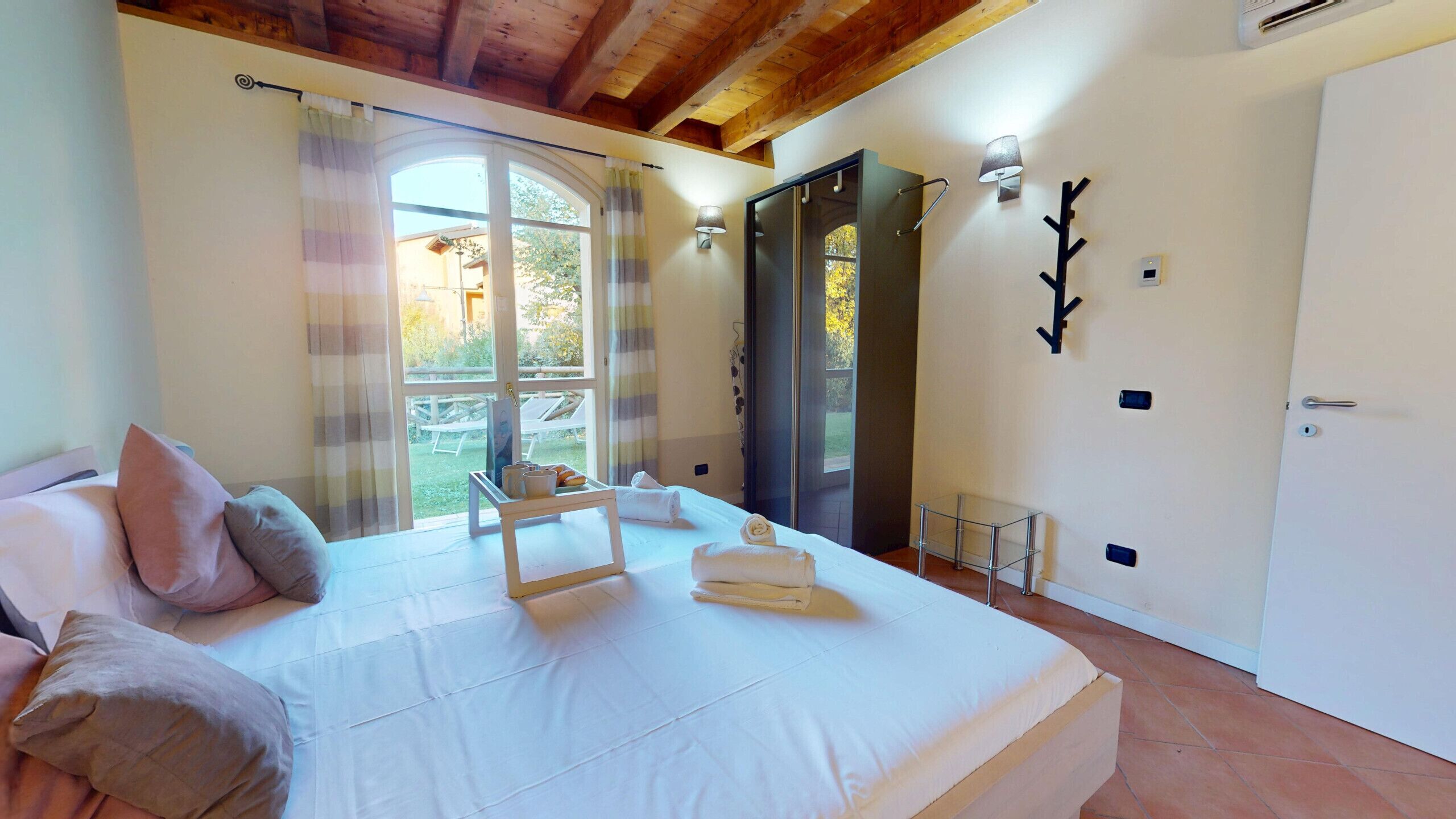 A bedroom with a large bed, a window with curtains, a door, and a wall-mounted light fixture.