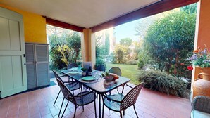 A covered patio with a table set for dining, surrounded by chairs, and a view of a garden with various plants and a wooden fence. - Borgo Torchio F1 By Bookinggardalake (Manerba del Garda)