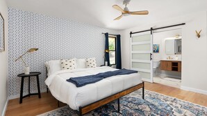 Une chambre à coucher avec un lit, un ventilateur de plafond, une table de chevet, une lampe, une fenêtre avec des rideaux, une salle de bain avec un miroir et un tapis à motifs.
