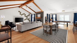 Un salon spacieux avec un plafond haut, des poutres apparentes, un canapé gris, une table à manger en bois avec des tabourets de bar, un téléviseur à écran plat et une cheminée.