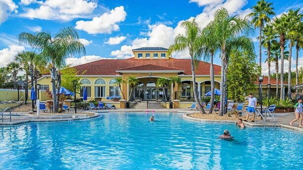 A swimming pool with palm trees, a building with a red roof, and a clear sky. - Lakefront Terra Verde Retreat Near Disney (Kissimmee)
