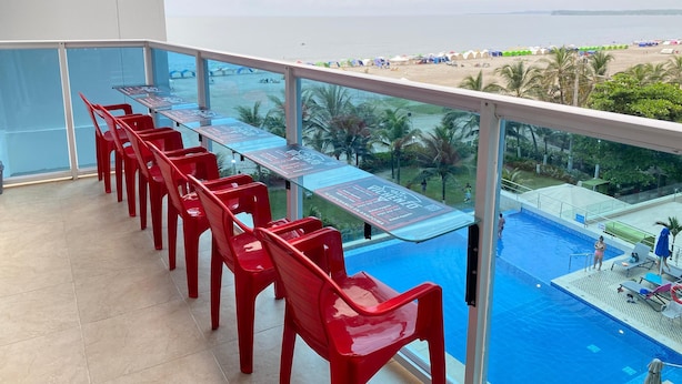 En balkon med røde stole og et glasbord med udsigt over en swimmingpool og strand.