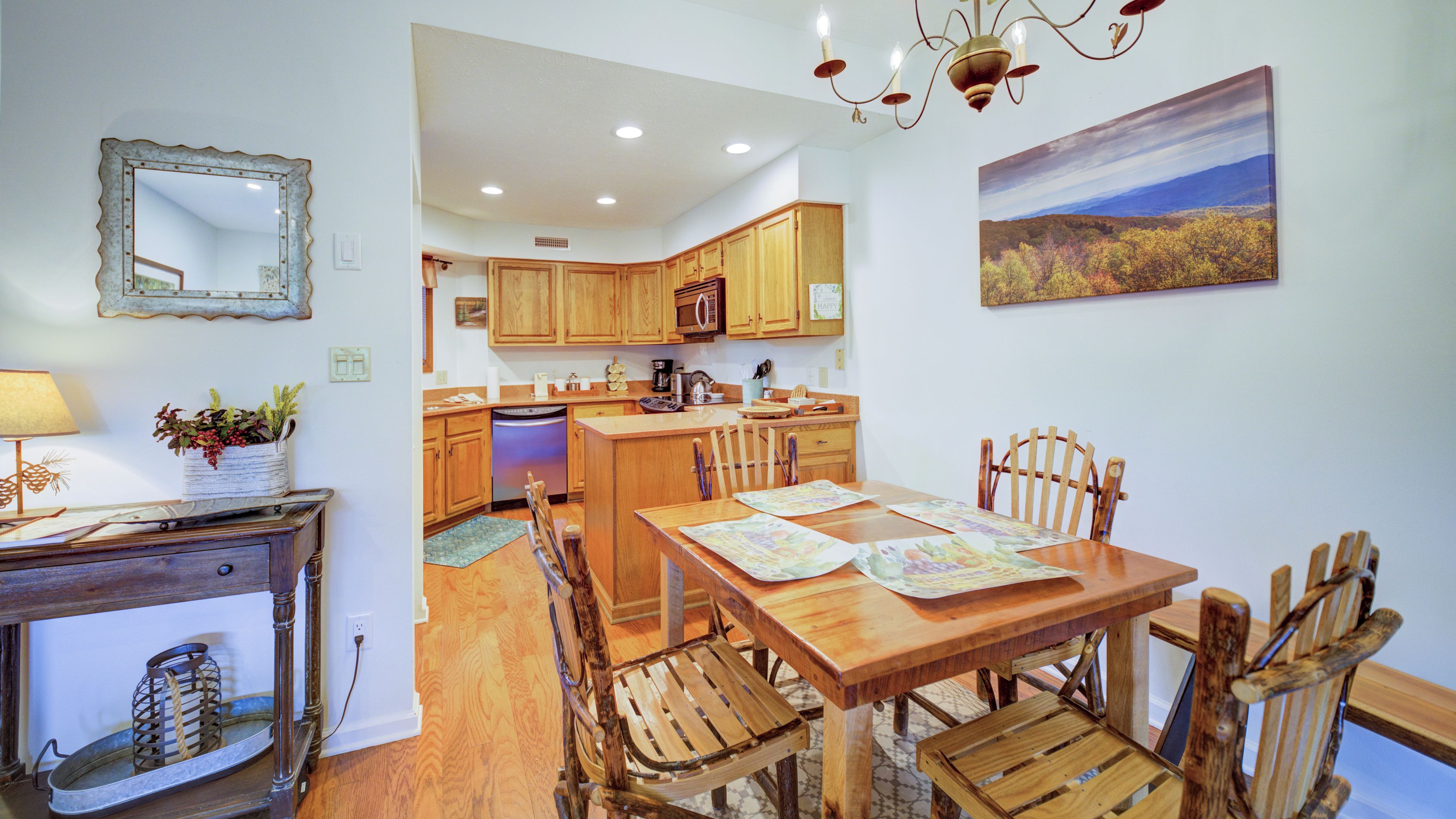 Une cuisine avec des meubles de rangement en bois, une table à manger avec des chaises, un lustre et un tableau représentant un paysage accroché au mur.