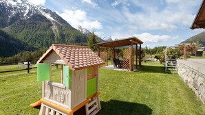 Una casita de juegos de madera con puerta verde y techo marrón, una pérgola de madera y una montaña al fondo.