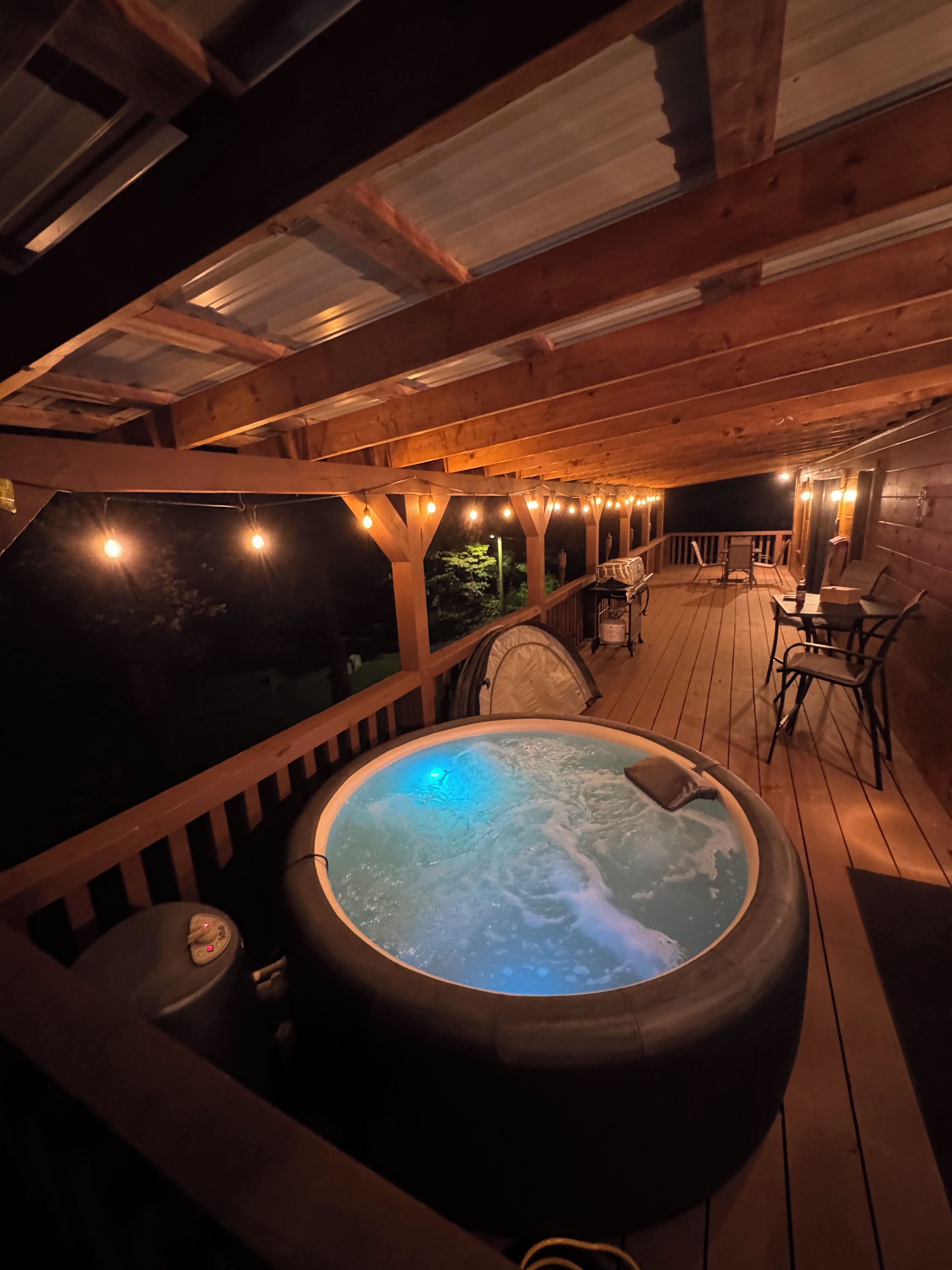 A hot tub on a wooden deck with string lights, surrounded by chairs and a table.