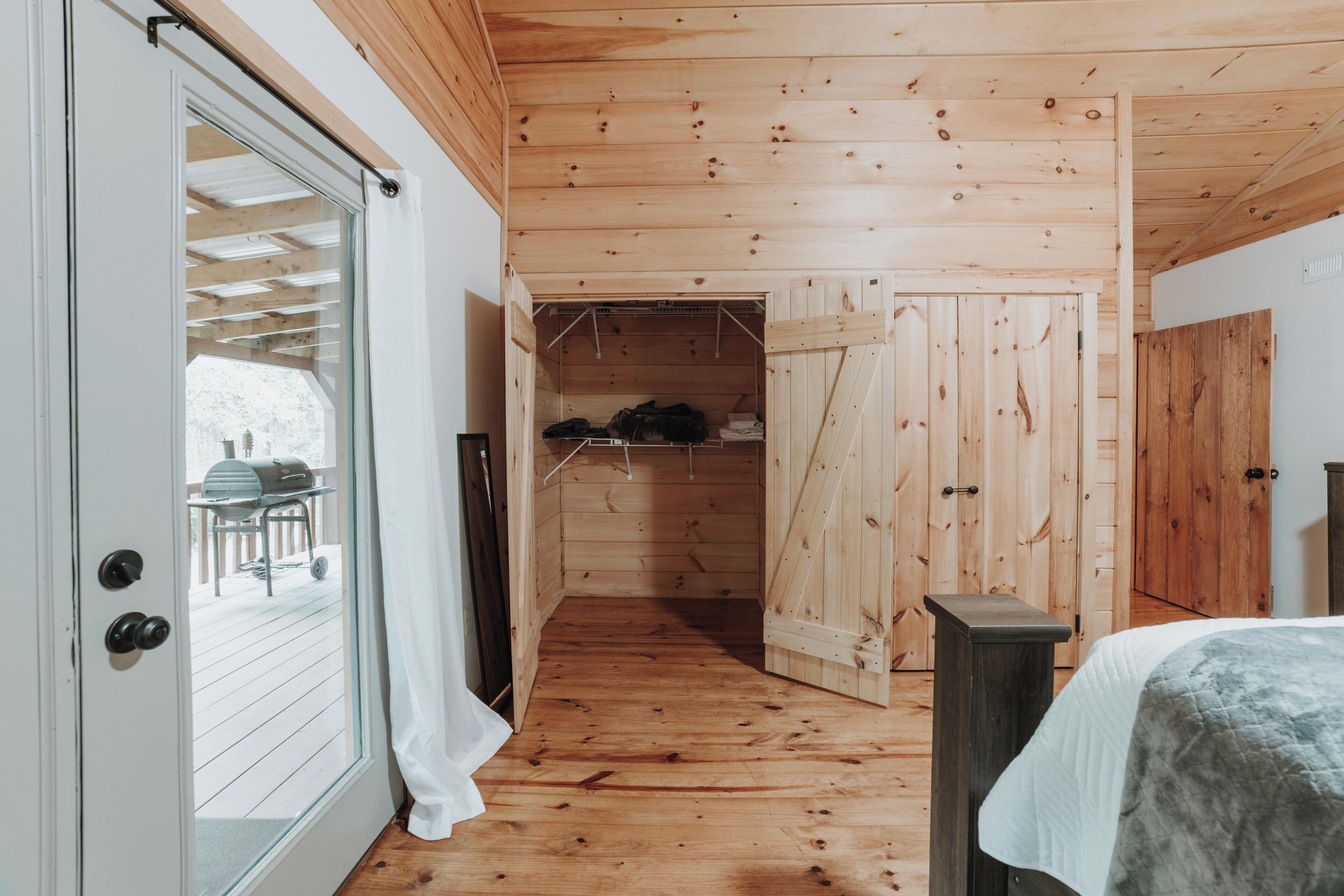 A wooden interior with a sliding door leading to an outdoor area, a bed with a white cover, and a wooden wardrobe.