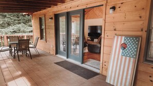 A wooden cabin with a covered porch, sliding glass doors leading to a living area with a TV and seating, and a flag on the wall. - Cozy 3-bedroom cabin with a spacious wrap around deck located in landisburg pa (Landisburg)