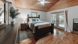 A bedroom with a wooden ceiling, a bed with a headboard, a nightstand, a bedside table, a potted plant, and a window with curtains. - Cozy 3-bedroom cabin with a spacious wrap around deck located in landisburg pa (Landisburg)