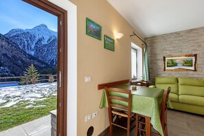 Een zithoek met een tafel en stoelen, een groene bank, uitzicht op een berg door een glazen deur, en een open haard met een lijst eromheen.