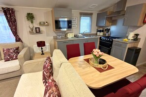 Een knus woongedeelte met een kitchenette, een bank, een eettafel met een bloemenvaas en een rode stoel.