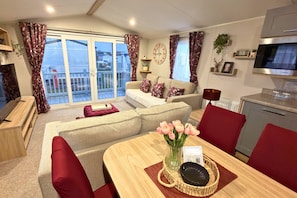 Een woonkamer met een bank, een eettafel met een bloemenvaas, een televisie en een keukenhoek.