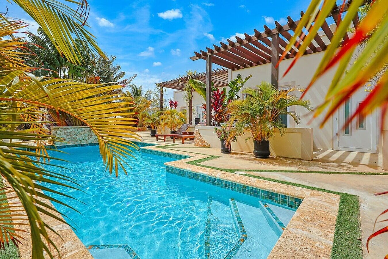 A swimming pool with tiled edges, surrounded by potted plants and a pergola.