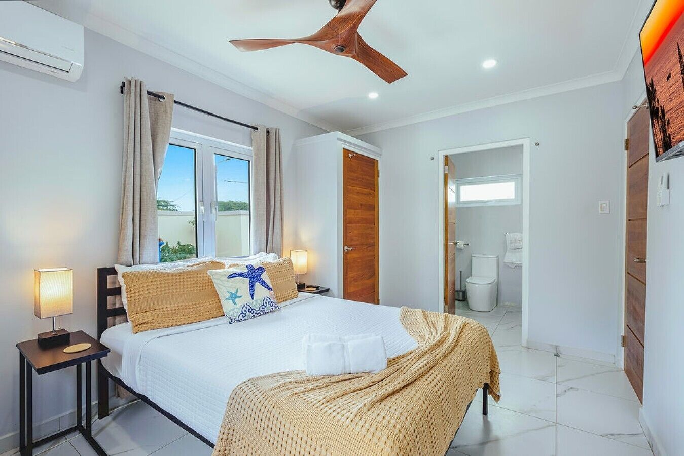 A hotel room with a bed, bedside tables, a ceiling fan, a window with curtains, and a view of the outdoors.
