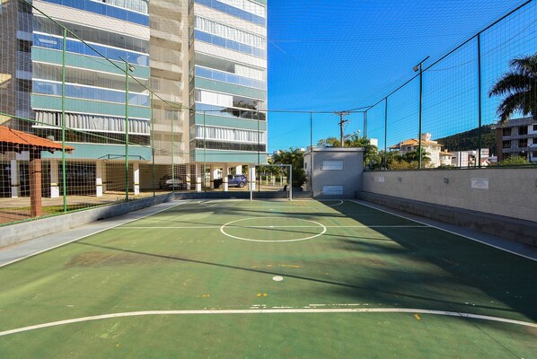 Una cancha de baloncesto al aire libre con superficie verde, rodeada por una valla y edificios.