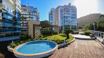 A rooftop pool area with a wooden deck, surrounded by greenery and modern buildings.
