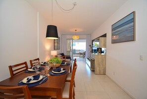 A dining area with a wooden table set for four, a hanging light fixture, and a view into the kitchen. - Boulevard Praia de Palmas a 400m do Mar (Governador Celso Ramos)
