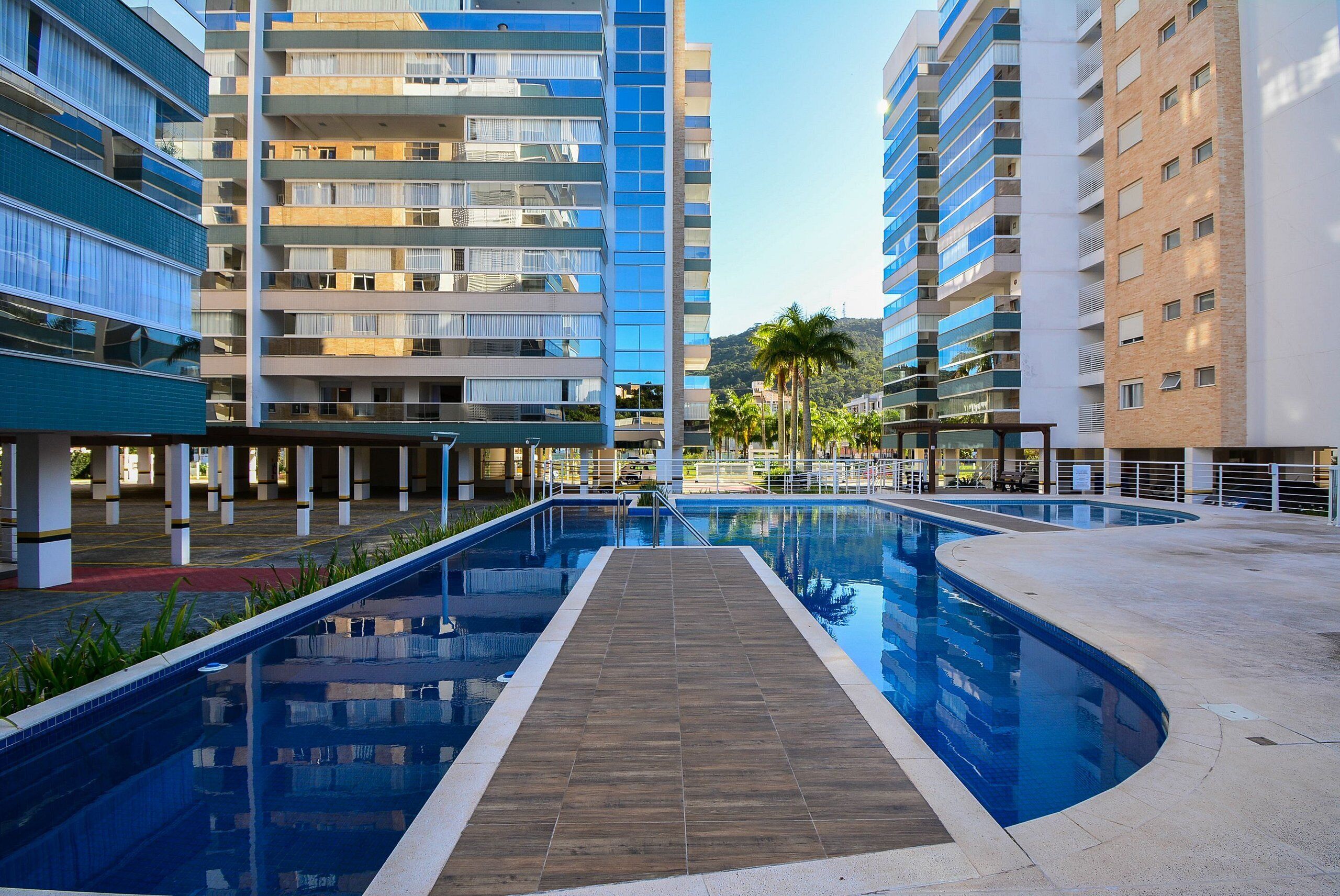 A modern hotel with a central swimming pool, multiple balconies, and a view of palm trees and hills.