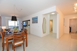 Un comedor moderno con una mesa de madera preparada para cuatro personas, una lámpara colgante y una cocina al fondo.