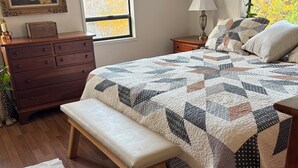 Een slaapkamer met een houten dressoir, een bankje, een geborduurde spreideken, een nachtkastje met een lamp, en een raam met uitzicht op bomen.
