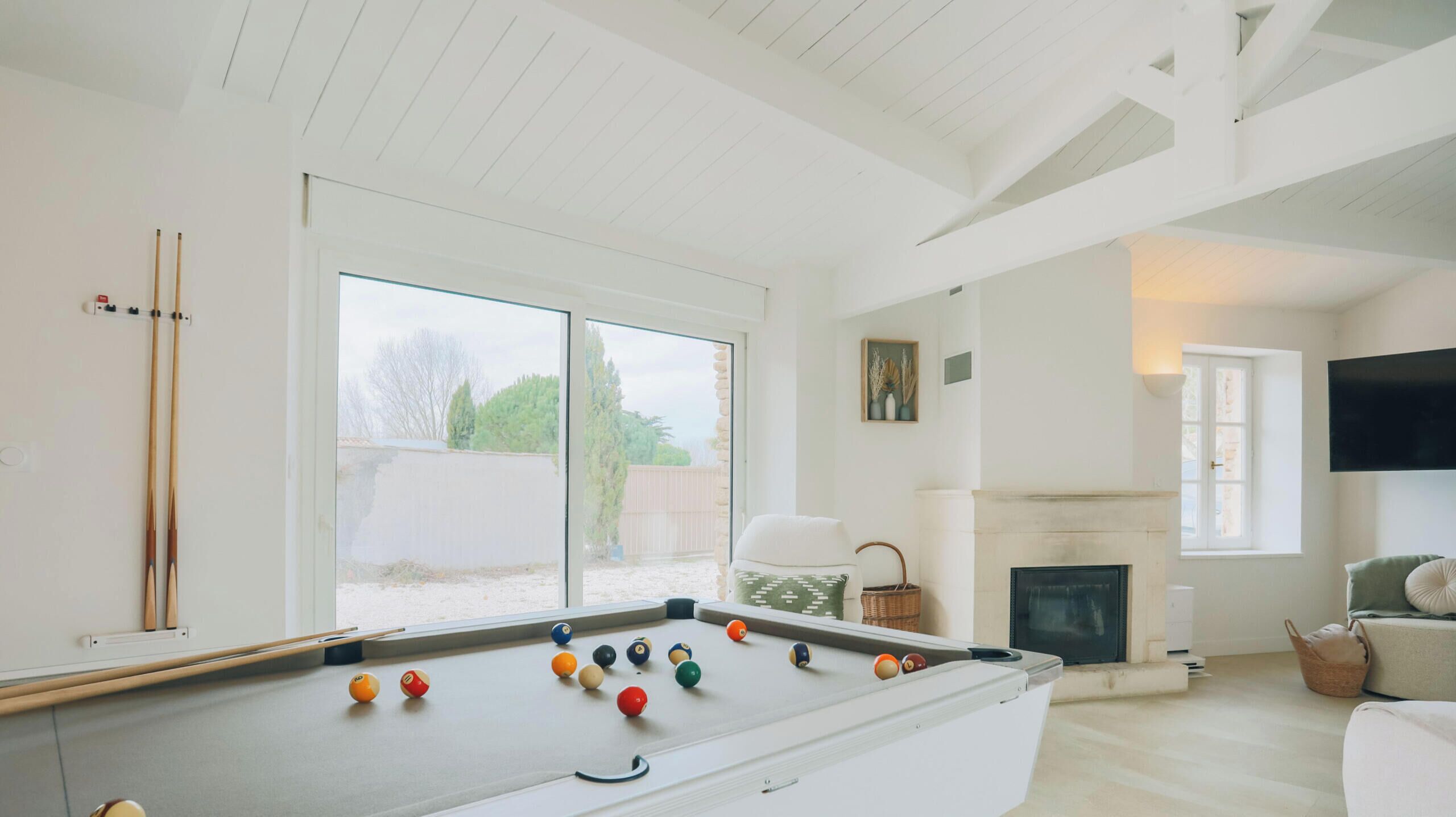 A pool table with balls, a sofa, a fireplace, and a TV in a bright room with white walls and wooden beams.