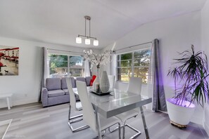 Un salon moderne avec une table à manger en verre, un canapé gris et une grande fenêtre agrémentée de rideaux.