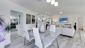 Uma área de refeições moderna e bem iluminada, com mesa de vidro, cadeiras brancas e um vaso decorativo.