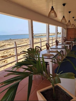 A seaside restaurant with outdoor seating, a view of the beach, and hanging pendant lights.