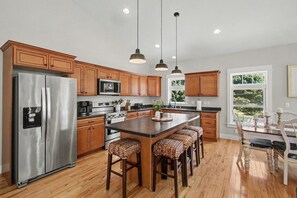 A modern kitchen with wooden cabinets, stainless steel appliances, a central island with stools, pendant lighting, and a view of the outdoors through a window. - 5-Min to South Beach | Golf Cart, Hot Tub & Arcade (South Haven)