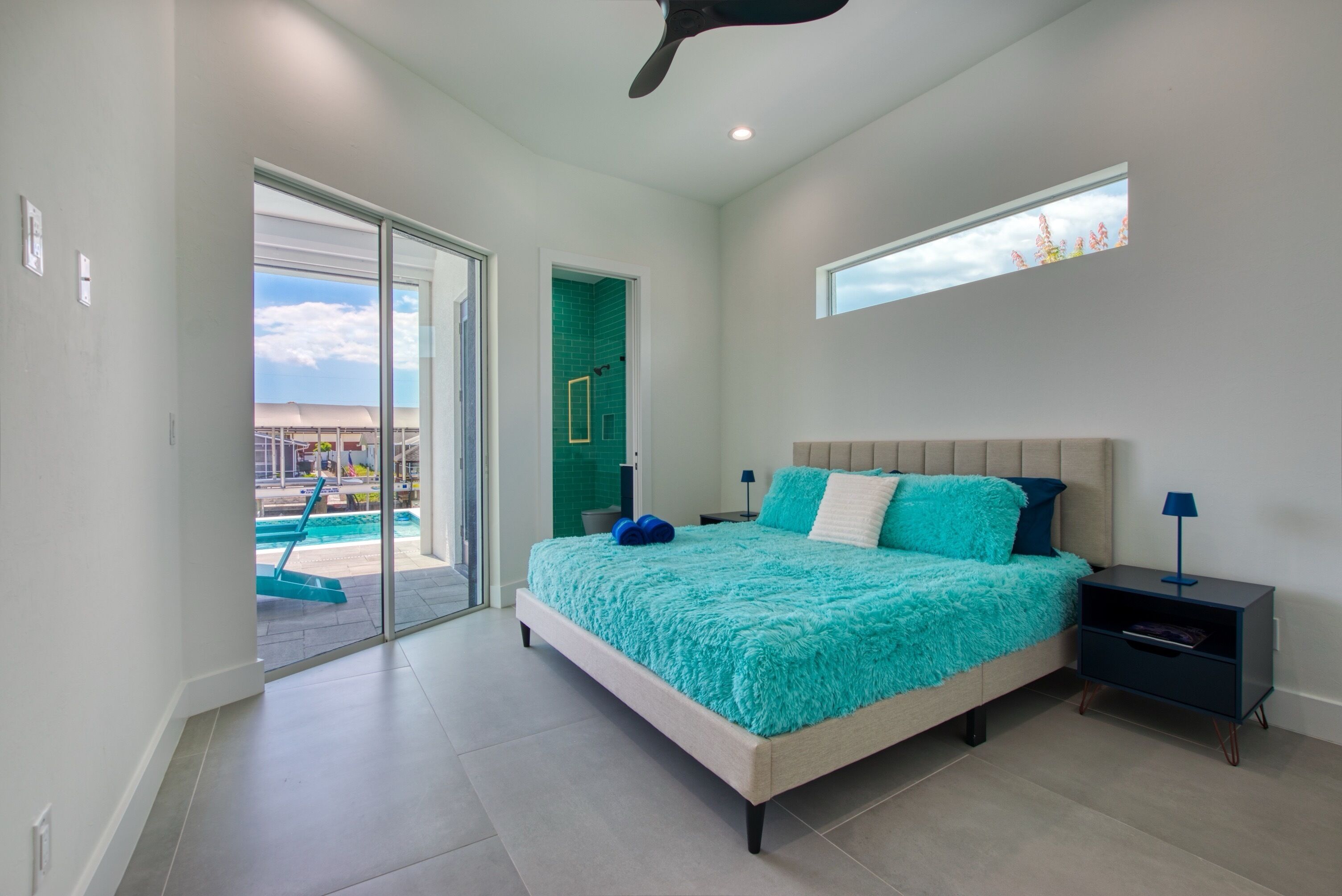 A modern bedroom with a large bed, a nightstand, and a view of a swimming pool through a glass door.