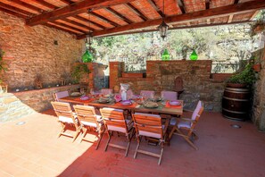 Un patio al aire libre con un muro de piedra, sillas de madera, una mesa lista para comer, faroles colgantes y plantas en macetas.