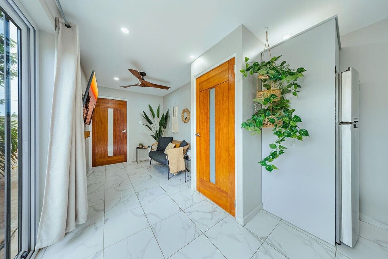 A modern interior with a hallway leading to a room, a ceiling fan, a door with a plant, and a window with curtains.