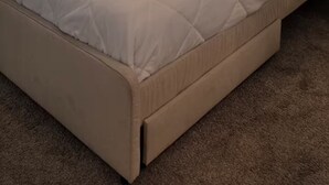 Ein ordentlich bezogenes Bett mit einer beigen Tagesdecke und zwei gefalteten Handtüchern, ein Fenster mit Jalousien und eine Nachttischlampe.