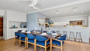 A modern dining area with a wooden table, blue upholstered chairs, and a kitchen in the background. - Somerset - Portsea - Coastal Escape with Private Beach Access (Portsea)