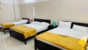 A hotel room with three beds, each with a black frame and white bedding. There are yellow throw blankets on each bed. The room has a tiled floor and a large window with blinds.