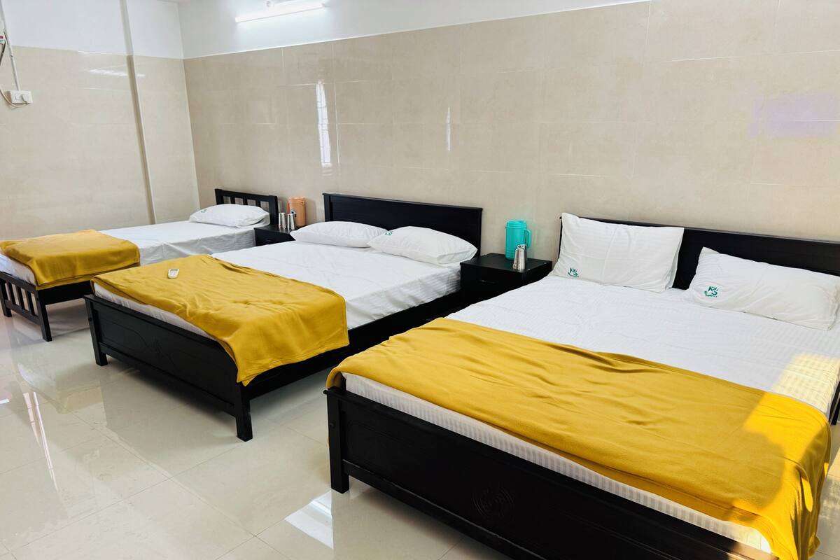 A hotel room with three beds, each with a black frame and white bedding. There are yellow throw blankets on each bed. The room has a tiled floor and a large window with blinds.